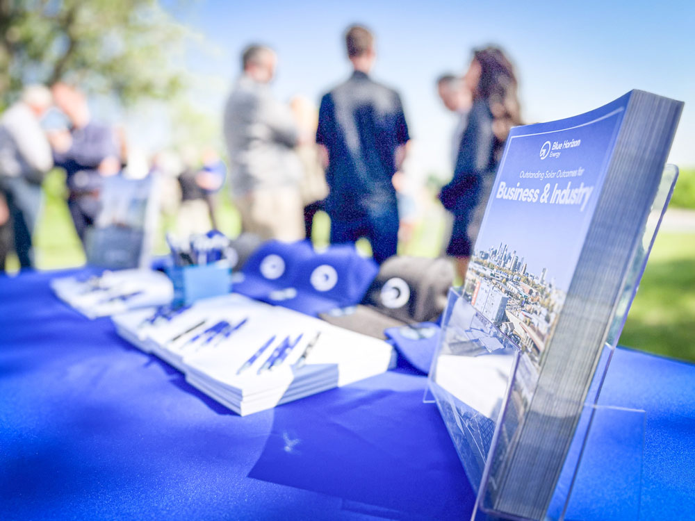 A collection of printed material and hats sit on a table with a crowd gathered in the background for the ribbon cutting event.